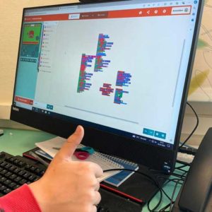 Computerbildschirm mit der Programmiersprache Scratch. Davor eine Kinderhand mit Daumen hoch.