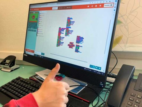 Computerbildschirm mit der Programmiersprache Scratch. Davor eine Kinderhand mit Daumen hoch.