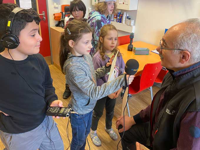 Mann sitzt und gibt drei Kindern, die vor ihm stehen, ein Interview. Ein Mädchen hält ihm das Mikrofon hin und einen Zettel in der Hand. Der Junge neben ihr guckt aufmerksam, hat Kopfhörer auf und das Aufnahmegerät in der Hand.