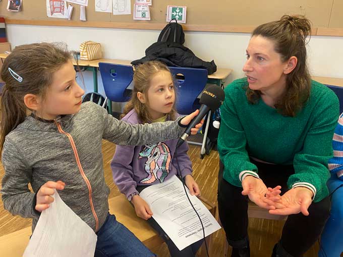 Frau in grünem Pullover sitzt im Klassenzimmer neben zwei Mädchen. Eins der Mädchen hält ihr das Radiofüchse-Mikrofon hin. Beide Mädchen haben Zettel mit Fragen in den Händen.