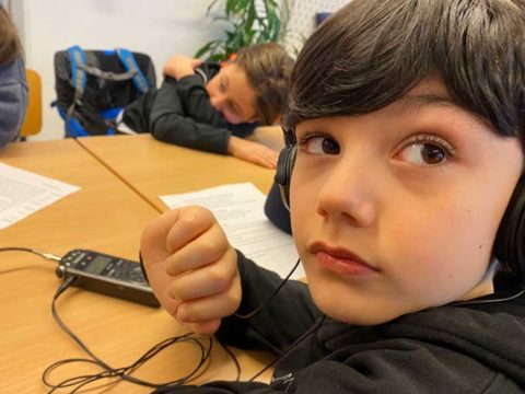 Junge im Vordergrund an einem Tisch hat Kopfhörer auf. Vor ihm liegt auf dem Tisch das Aufnahmegerät.