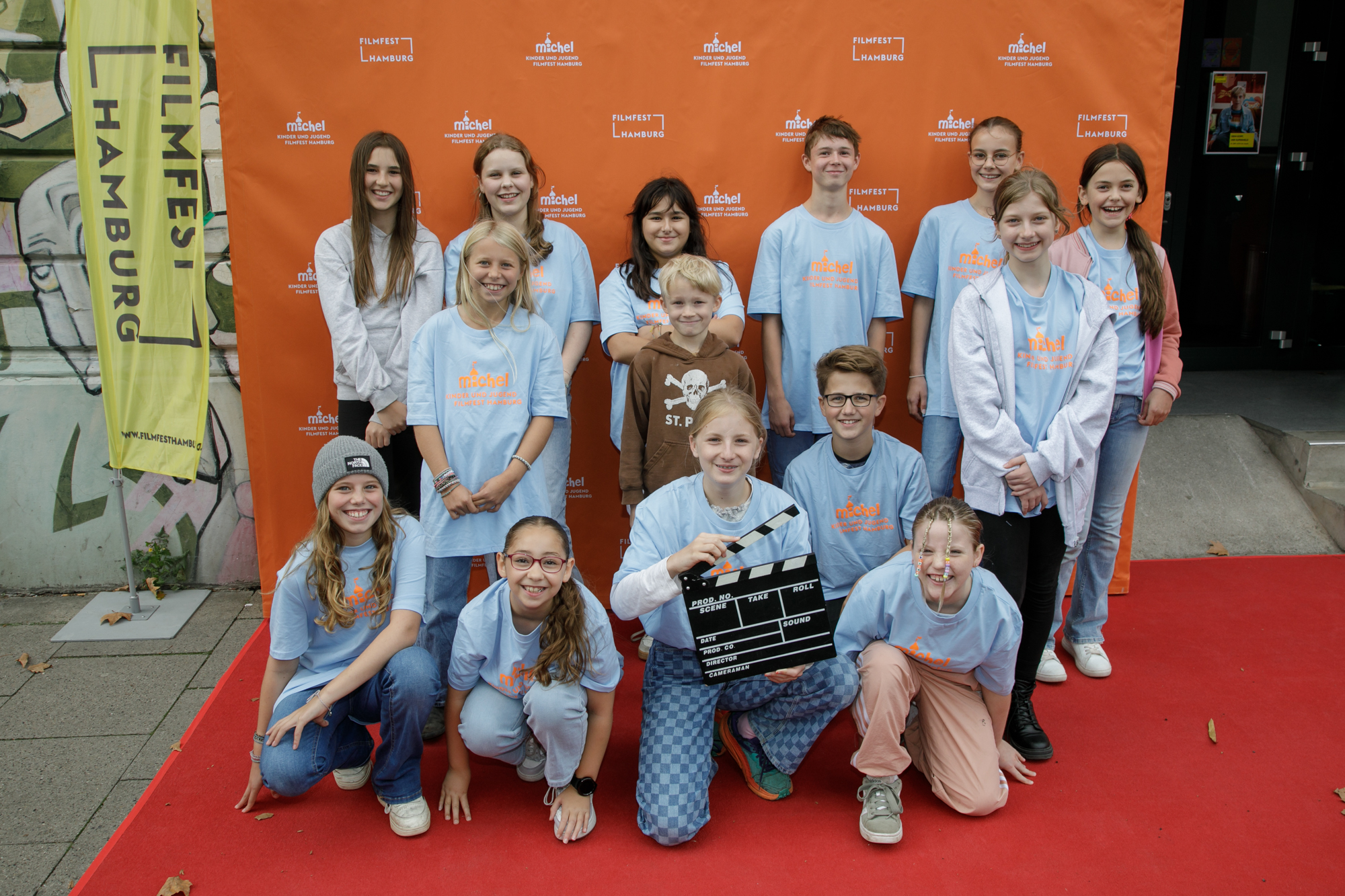 Vierzehn Kinder stehen und sitzen auf einem roten Teppich vor einer orangenen Wand und lächeln in die Kamera. 