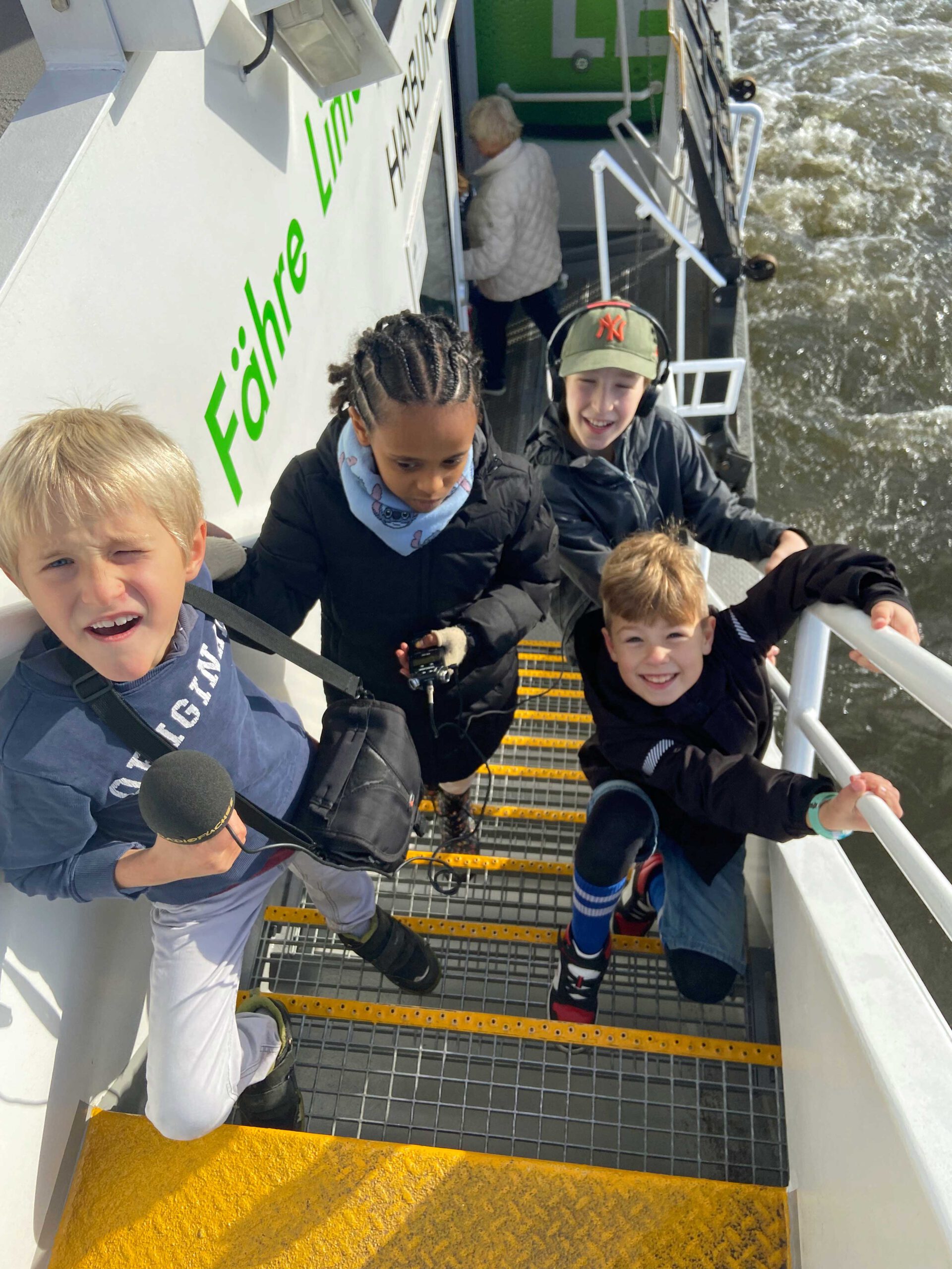 Vier Kinder gehen mit Aufnahmegerät und Mikrofon die Treppe einer Fähre hoch.