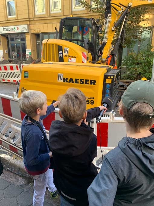 Drei Jungs stehen vor einem Bagger und halten das Radiofüchse Mikrofon hin