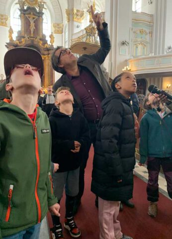 Kinder stehen mit einem Mann in einer Kirche und gucken nach oben, vor ihnen ein Aufnahmegerät